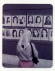 Madre con pañuelo, observando fotocopias con imágenes de mujeres desaparecidas sobre muro urbano. 
